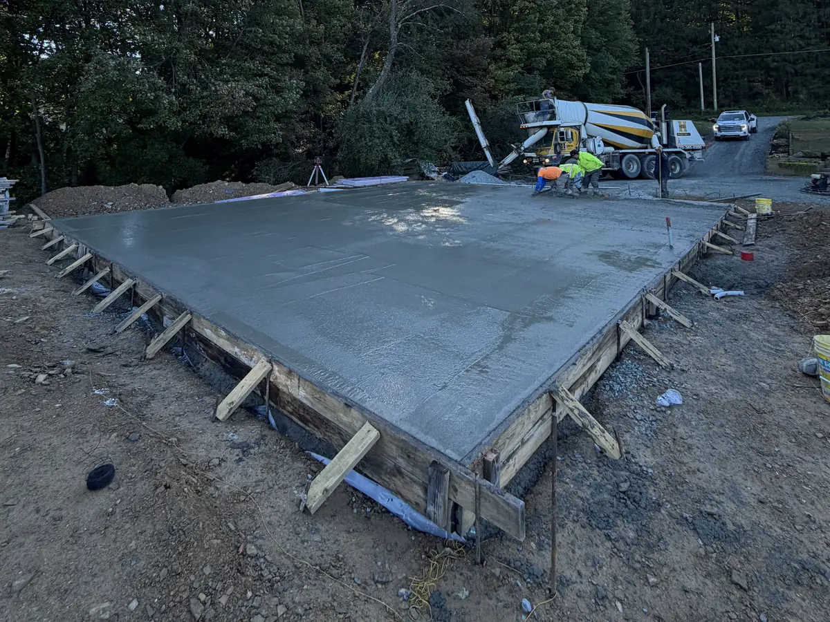 A slab and its timber forms surrounded by site ground, with a mixer truck and crew in the background